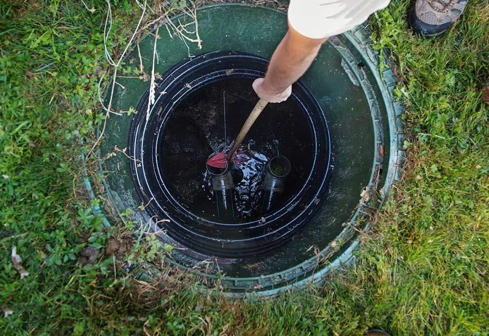A technician lowering a tool into an open septic tank riser to check sludge depth