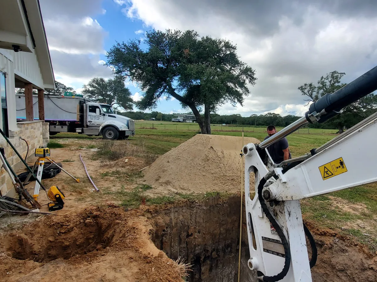An excavator digging a septic tank pit beside a house on a rural lot
